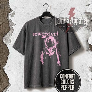 Puede incluir: Una camiseta de manga corta de color gris carbón con un estampado gráfico rosa de un personaje de estilo anime. El texto "cyberia lyfe" y "sewerslvt" está impreso en rosa sobre el personaje. La camiseta cuelga de una percha negra.