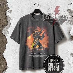 Może przedstawiać: Ciemnoszary t-shirt z krótkim rękawem z nadrukiem graficznym przedstawiającym żołnierza w zbroi trzymającego dwie bronie pośród pomarańczowych i żółtych płomieni. Tekst Halo 2 jest wydrukowany pod grafiką wraz ze zdaniem There are those who said this day would never come. What are they to say now?