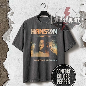 Puede incluir: Una camiseta de manga corta en color gris carbón con un gráfico de estilo vintage de la banda Hanson. El estampado incluye el texto HANSON, la frase THIS TIME AROUND y una imagen fotográfica de los tres integrantes en tonos cálidos.