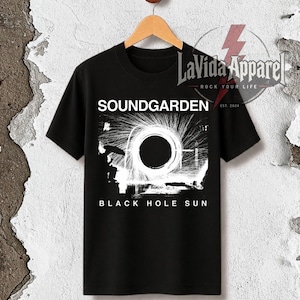 May include: A black short-sleeve t-shirt featuring a white graphic print of a circular light effect. The text SOUNDGARDEN appears above the graphic, and the text BLACK HOLE SUN appears below it. A logo for LaVida Apparel is visible in the top right.