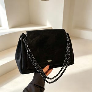 May include: A black handbag with a soft matte texture, featuring a fold-over flap and a dual-chain shoulder strap woven with black material. The front of the flap displays small gold-toned lettering that reads FASHION&BAGS LEATHER.