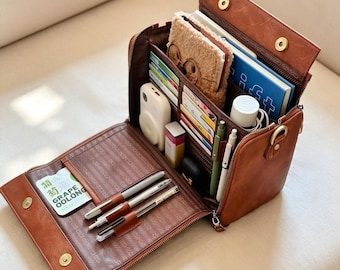 May include: A brown leather-effect organiser bag with a fold-out flap featuring multiple interior slots and pockets. It contains pens, a small white camera, a notebook with a fuzzy cover, a blue book, and a card labelled Grape Oolong.