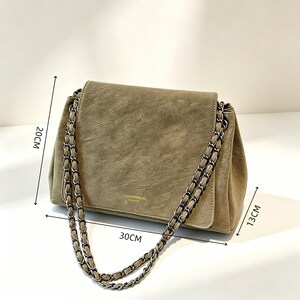 May include: An olive green shoulder bag with a textured finish and a dark metal chain strap. The bag features a flap closure with small gold text. Dimensions are indicated as 20 cm in height, 30 cm in width, and 13 cm in depth by arrows.