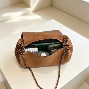 May include: A tan shoulder bag with a flap closure and a dark metal chain strap. The interior is open and contains a green smartphone, a small black and white striped rectangular item, a grey bottle, and a small zippered pouch inside.