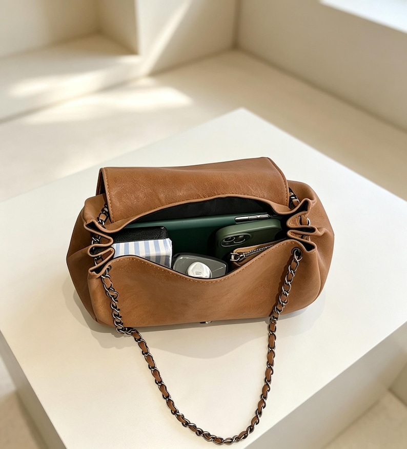 May include: A tan shoulder bag with a flap closure and a dark metal chain strap. The interior is open and contains a green smartphone, a small black and white striped rectangular item, a grey bottle, and a small zippered pouch inside.