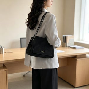 May include: A black shoulder bag with a suede-like texture, featuring a rectangular flap, a gold-toned metal logo plate, and a chain-link strap interwoven with black material. The bag is worn over the shoulder, showcasing its structured design.