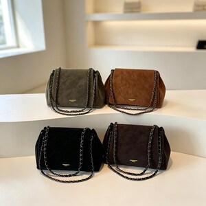 May include: Four rectangular shoulder bags with a soft, suede-like texture, each featuring a gold-toned metal chain strap and a small gold logo plaque on the front. The bags are available in olive green, tan, black, and dark brown colour variations.