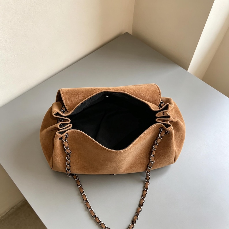 May include: A tan suede shoulder bag with a soft, slouchy silhouette and a fold-over flap. The bag features a dark metal chain strap woven with matching tan suede and a black fabric interior lining, visible through the open top compartment.