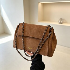 May include: A brown shoulder bag with a textured finish, featuring a fold-over flap and a dark metal chain strap woven with matching brown material. The front of the bag displays small, subtle gold-toned text centred near the bottom edge.
