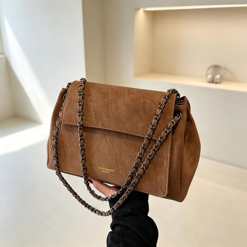 May include: A brown shoulder bag with a textured finish, featuring a fold-over flap and a dark metal chain strap woven with matching brown material. The front of the bag displays small, subtle gold-toned text centred near the bottom edge.