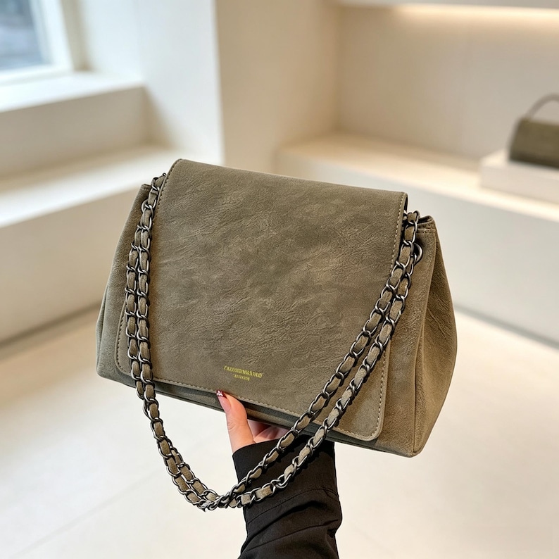 May include: A sage green shoulder bag with a textured finish and a flap closure. The bag features a dark metallic chain strap woven with matching green material. A small, rectangular gold-toned logo plate is centred on the lower front flap.