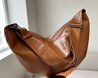 May include: A crescent-shaped brown shoulder bag with a smooth, slightly glossy finish. The bag features a wide, adjustable strap connected by dark metal hardware, a top zip closure, and visible stitched paneling along the curved body.