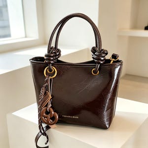 Peut inclure: Un sac à main marron foncé, brillant et texturé, doté de deux anses nouées et de détails dorés. Une breloque décorative composée d'un petit boulier et d'un pendentif en forme de bretzel est suspendue sur le côté. L'inscription FASHION & BAGS est visible.