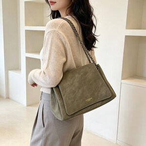 May include: An olive green shoulder bag with a soft, suede-like texture, featuring a rectangular flap closure, a gold-toned metal logo plate, and a dark chain-link strap that rests over the shoulder for a structured and functional accessory.