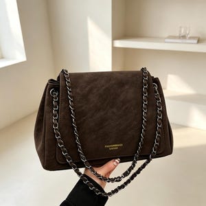 May include: A dark brown shoulder bag with a textured finish and a flap closure. The bag features a long silver-toned metal chain strap interwoven with matching brown material. The text FASHIONBBAGS LEATHER is printed in small gold letters on the front.
