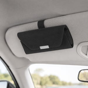 May include: A black rectangular car visor sunglasses holder with a fold-over flap and a small silver-toned rectangular accent piece on the front. The holder is secured to the light gray vehicle sun visor by a black strap attached to the back.