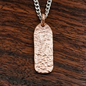 May include: A rose gold-toned rectangular pendant with a rounded top and a hammered, textured surface. The pendant is suspended from a silver-toned metal chain link necklace. The metal surface reflects light across its uneven, dimpled finish.