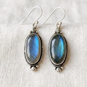Peut inclure: Une paire de boucles d'oreilles pendantes en argent avec des pierres de labradorite ovales aux reflets bleus. Les pierres sont serties dans des montures en argent avec des bordures torsadées et de petites perles décoratives en haut et en bas.