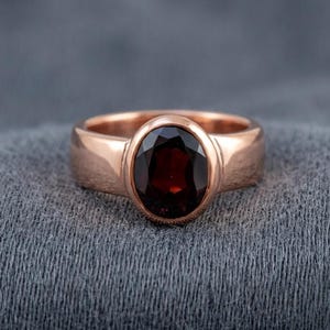 May include: A rose gold ring featuring a large, oval-cut, deep red gemstone set in a smooth, polished band. The dark red stone is faceted and held securely in a bezel setting, creating a clean, continuous look for the metal band surrounding it.
