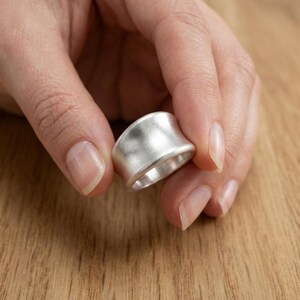 Può includere: Un ampio anello in argento con una finitura liscia e opaca e una superficie leggermente concava e curva. La fascia presenta una solida struttura metallica con una lucentezza uniforme e tenue su tutto l'esterno, mostrando una forma arrotondata.