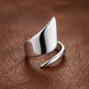 Puede incluir: Un anillo de tono plateado pulido con un diseño abierto de estilo bypass, que presenta una banda de metal lisa, curva y cónica que se envuelve alrededor del dedo. La superficie es altamente reflectante, mostrando un acabado metálico sólido.