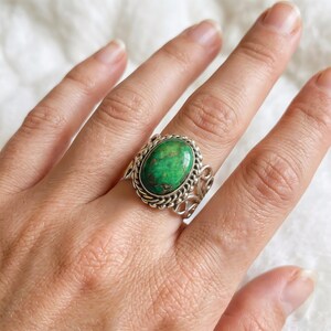 May include: A silver ring featuring a large, oval-shaped green stone with dark brown matrix veining. The stone is set in a twisted rope border, and the band is crafted with an openwork design consisting of swirling metal patterns.
