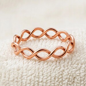 Op de afbeelding: Een roségoudkleurige ring met een doorlopend gedraaid draadontwerp. De band is gemaakt van een enkele metalen streng die herhaaldelijk lussen vormt om een open, onderling verbonden patroon rond de gehele omtrek van de ring te creëren.