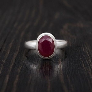 Op de afbeelding: Een zilveren ring met een centrale, ovaalvormige, gefacetteerde dieprode edelsteen. De steen is gezet in een gladde, gepolijste zilveren zetting en de ringband is van massief, effen zilvermetaal met een constante breedte rondom de vinger.