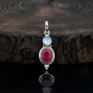May include: A silver pendant featuring a faceted oval ruby gemstone set below a smaller, round, translucent moonstone. The pendant has a polished silver frame with a small decorative bead detail at the bottom and a silver bail at the top.