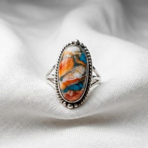 May include: A silver ring featuring a large, oval-shaped stone with a marbled pattern of vibrant orange, bright turquoise, white, and metallic grey veins. The stone is set in a polished silver bezel surrounded by a decorative twisted rope border.