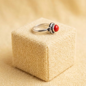 May include: A silver ring featuring a smooth, polished band and a central setting with a vibrant, round, red stone. The stone is encircled by a decorative, twisted rope-style metal border that adds texture to the circular focal point of the piece.