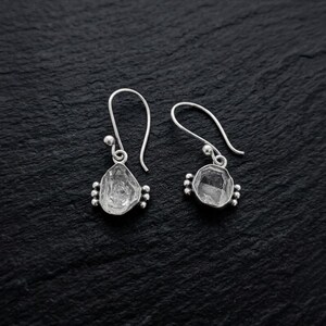 Peut inclure: Une paire de boucles d'oreilles pendantes en argent avec des pierres brutes, transparentes et de forme irrégulière serties dans des montures en métal. Chaque boucle possède un crochet incurvé et trois petites sphères décoratives en argent.