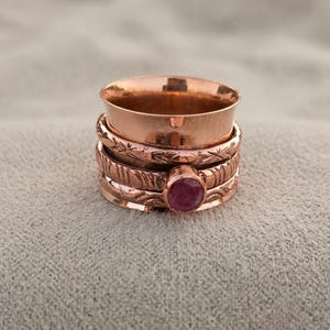 May include: A copper spinner ring featuring a wide, smooth band at the top, followed by a textured band with etched patterns, and a bottom band set with a single, round, dark pink gemstone. The metal has a warm, metallic, reddish-brown finish.
