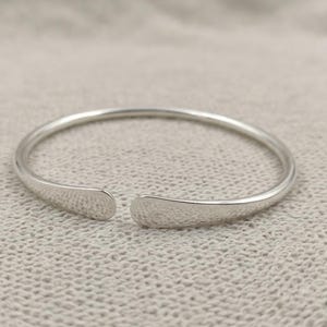 May include: A silver-coloured open cuff bracelet with a smooth, polished finish. The bracelet features a circular band that tapers slightly towards the two rounded, open ends, creating a simple and continuous metallic loop design.