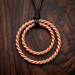 Peut inclure: Un pendentif composé de deux cercles concentriques en fil de cuivre torsadé. Les cercles sont suspendus à un cordon noir noué sur le dessus, créant un design circulaire superposé avec une finition en cuivre métallique et un cordon sombre.