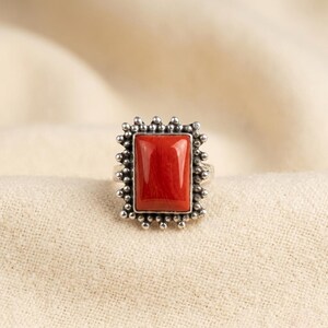May include: A silver ring featuring a rectangular, smooth, vibrant red stone set in the center. The setting is surrounded by a decorative border of small, raised silver beads, creating a textured frame around the polished red gemstone.