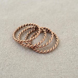 Op de afbeelding: Een set van drie dunne, ronde ringen van koperkleurig metaal. Elke ring heeft een doorlopende gedraaide touwtextuur die een herhalend spiraalpatroon rond de gehele omtrek van de band creëert, met een metallic afwerking.