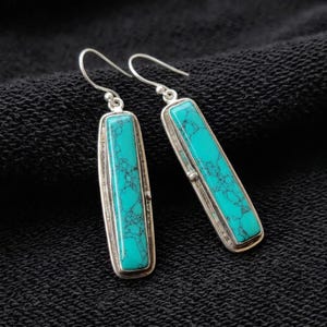 May include: A pair of drop earrings featuring long, rectangular turquoise stones with dark matrix veining, set in polished silver frames. Each earring hangs from a simple silver hook, showcasing a sleek, elongated design with vibrant blue-green colour.