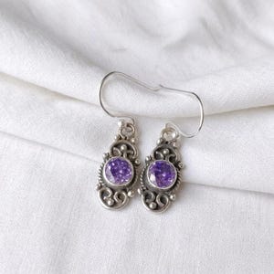 May include: A pair of drop earrings featuring round, faceted purple stones set in silver-toned metal. The settings display intricate, swirling wirework patterns and small bead accents. Each earring hangs from a simple, curved silver-toned ear wire.