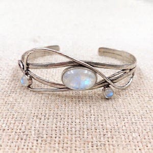 Può includere: Un bracciale rigido in argento con una grande pietra di luna ovale al centro e due pietre di luna rotonde più piccole ai lati. La fascia in metallo è composta da diversi fili d'argento sottili, sovrapposti e curvi dal design aperto.