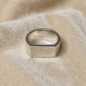 Peut inclure: Une bague de couleur argentée présentant une large face rectangulaire et plate qui se prolonge par un anneau lisse et arrondi. La surface métallique possède une finition polie et réfléchissante, offrant un aspect uniforme et épuré.