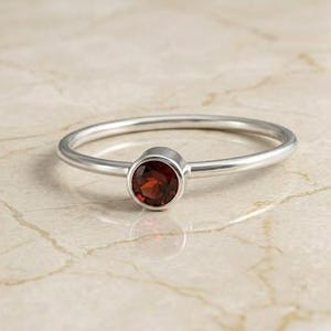 Pode incluir: Um anel fino de prata com uma única pedra preciosa granada redonda de cor vermelho profundo, montada num engaste liso. O aro é polido e fino, realçando o contraste entre o acabamento metálico prateado e a pedra de cor vermelho escuro.