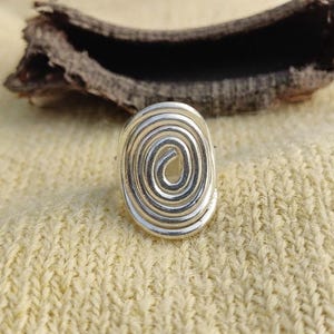 May include: A silver-toned ring featuring a prominent, raised spiral design on its face. The metal surface is smooth and reflective, forming a continuous coiled pattern that creates an oval shape. The ring is constructed from a single piece of wire.