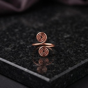 Könnte beinhalten: Ein kupferfarbener Ring mit offenem Banddesign und zwei symmetrischen Spiralmotiven an den Enden. Der Metalldraht ist zu engen, kreisförmigen Mustern gewickelt, die nach außen zeigen und eine durchgehende, fließende Form für den Finger bilden.