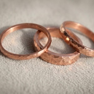 Puede incluir: Tres anillos de color cobre con una textura martillada distintiva en la superficie exterior. Los anillos varían ligeramente en ancho y están dispuestos en un grupo superpuesto, mostrando el brillo metálico y el acabado facetado irregular.