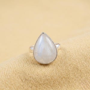 May include: A silver ring featuring a large, pear-shaped moonstone cabochon set in a smooth metal bezel. The gemstone displays a translucent, milky white appearance with subtle iridescent blue flashes, mounted on a simple, polished silver band.