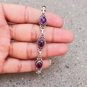 May include: A silver bracelet featuring three oval-shaped, deep purple amethyst gemstones. Each stone is set in a polished silver bezel with a twisted rope-style border, connected by small silver links to form a continuous chain design.