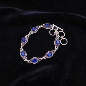May include: A silver bracelet featuring six oval-cut deep blue stones set in twisted metal frames. The bracelet is secured with a toggle clasp consisting of a T-bar and two circular rings, connected by small silver links between each stone setting.