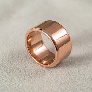 May include: A wide band ring with a polished rose gold finish. The ring features a smooth, reflective surface and a thick, solid construction with rounded edges, showcasing a metallic copper-toned hue throughout the entire circular band.