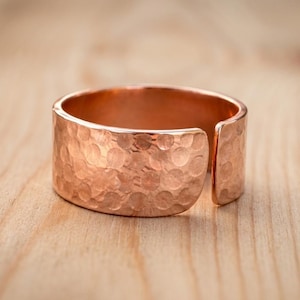 Può includere: Un anello largo e aperto realizzato in rame lucido. La superficie esterna presenta una distinta texture martellata, che crea una serie di piccole rientranze circolari lungo la fascia metallica. L'anello ha una finitura liscia e riflettente.